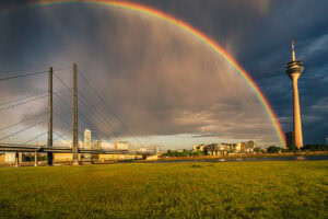 Fine-Art-Print Regenbogen über dem Rheinturm in Düsseldorf bei Unwetter – Panorama mit Rheinwiesen, Brücke und dramatischem Himmel, erhältlich als hochwertiger Kunstdruck.