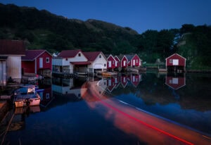 Lichtspuren eines Bootes bei Abenddämmerung vor roten Fischerhütten in Hidra, Norwegen – als exklusiver Fine Art Print erhältlich.