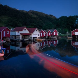 Lichtspuren eines Bootes bei Abenddämmerung vor roten Fischerhütten in Hidra, Norwegen – als exklusiver Fine Art Print erhältlich.