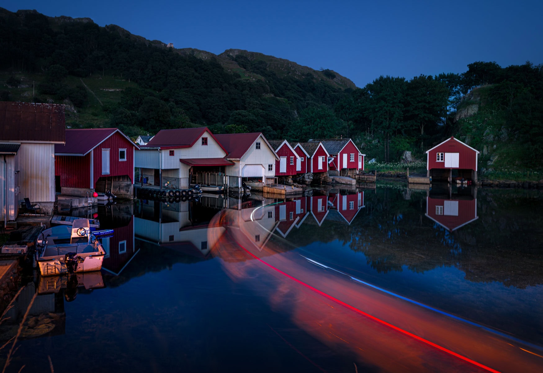 Lichtspuren eines Bootes bei Abenddämmerung vor roten Fischerhütten in Hidra, Norwegen – als exklusiver Fine Art Print erhältlich.