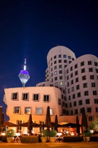 Fine Art Print der Gehry-Bauten in Düsseldorf bei Nacht mit leuchtendem Rheinturm und klar erkennbarem Sternenhimmel im Hintergrund.