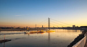 Fine Art Print: Sonnenaufgang über dem Rhein mit Frachtschiff, Rheinkniebrücke und Skyline von Düsseldorf in klarem Morgenlicht.