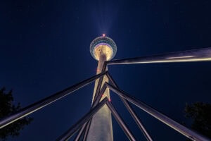 Fine Art Print des beleuchteten Rheinturms in Düsseldorf bei Nacht mit Sternenhimmel, aufgenommen aus einer ungewöhnlichen Perspektive.
