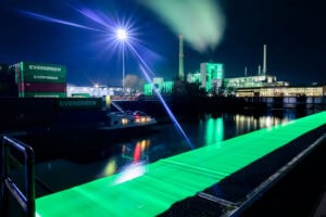 Düsseldorf Industriehafen Lausward – grünes Lichtband über dem Wasser, Laternen-Lichtstern und Rauchfahne, Nacht, Langzeitbelichtung.