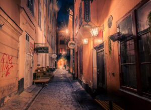 Nachtaufnahme enger Gasse in Gamla Stan, Stockholm, warmes Laternenlicht und historische Hausfassaden.