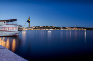 Weitwinkel-Nachtbild: Boot mit schwedischer Flagge links, weites Wasser und die Lichter von Djurgården/Stockholm im Hintergrund; sanfte Reflexionen.