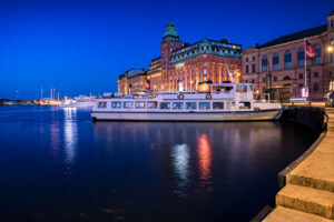 Weiße Fähre an Nybroviken vor dem Grand Hôtel in Stockholm bei blauer Stunde, ruhiges Wasser mit Lichtreflexionen.