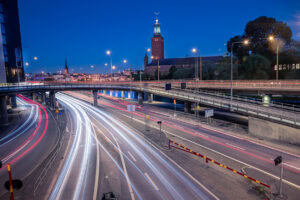 Stadtansicht mit Stockholms Stadshuset, Centralbron und hellen Autolichtspuren bei blauer Stunde; Laternen als Sterne, Mondsichel am Himmel.