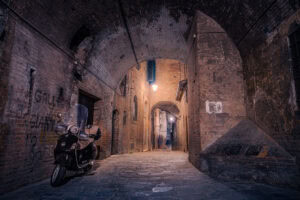 Siena, Toskana – enge Altstadtgasse bei Nacht mit Vespa und Laterne unter Backsteinbogen.