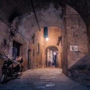 Siena, Toskana – enge Altstadtgasse bei Nacht mit Vespa und Laterne unter Backsteinbogen.