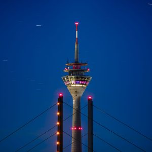 Beleuchteter Fernsehturm hinter Brückenpylonen vor tiefblauem Nachthimmel