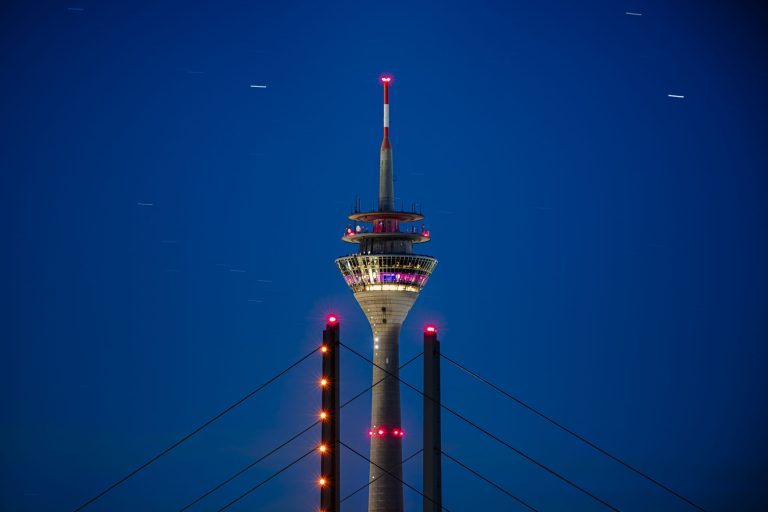 Beleuchteter Fernsehturm hinter Brückenpylonen vor tiefblauem Nachthimmel