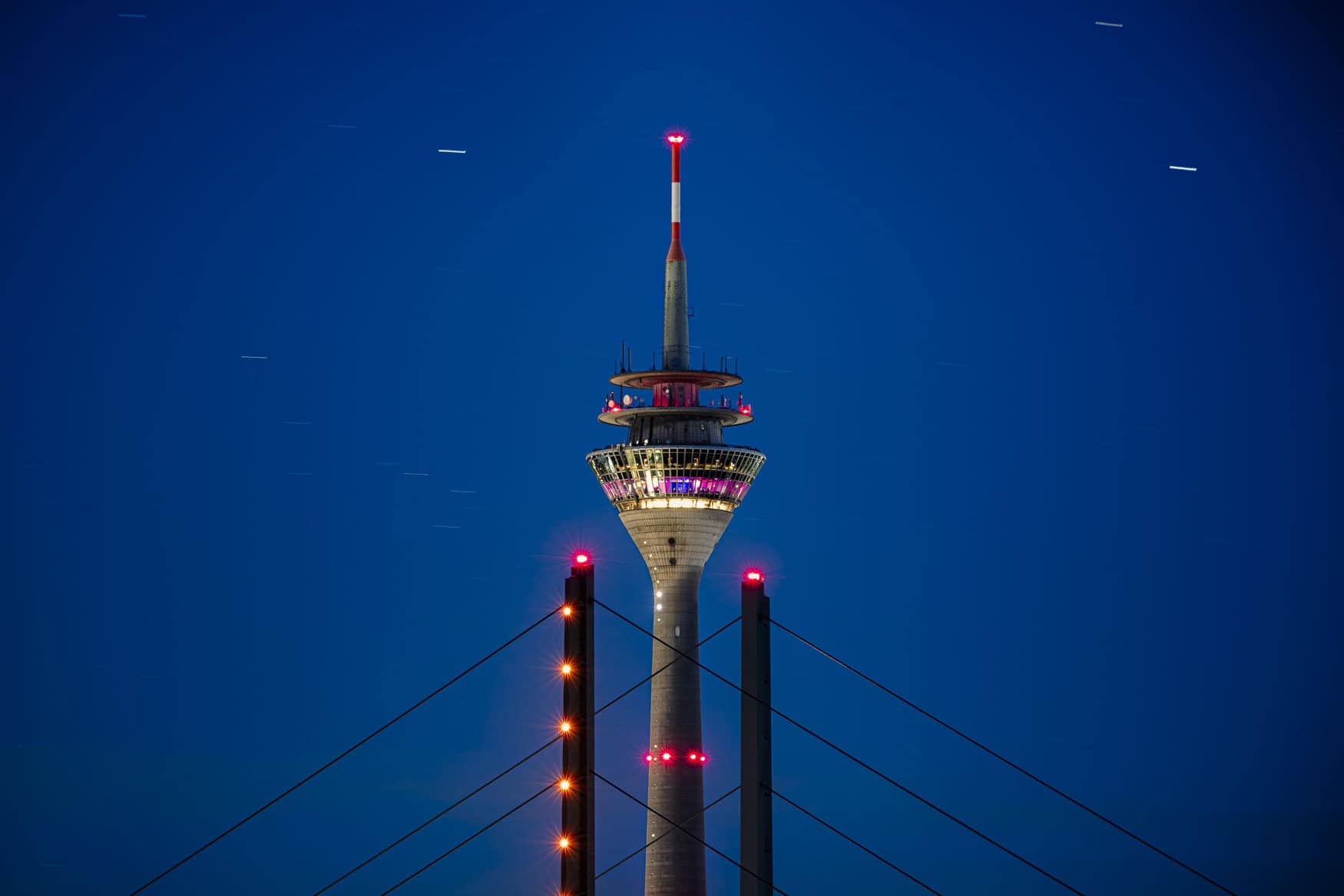 Beleuchteter Fernsehturm hinter Brückenpylonen vor tiefblauem Nachthimmel