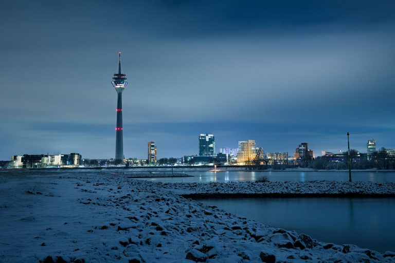 Verschneite Rheinuferwiesen mit Düsseldorfer Rheinturm und Skyline bei Nacht