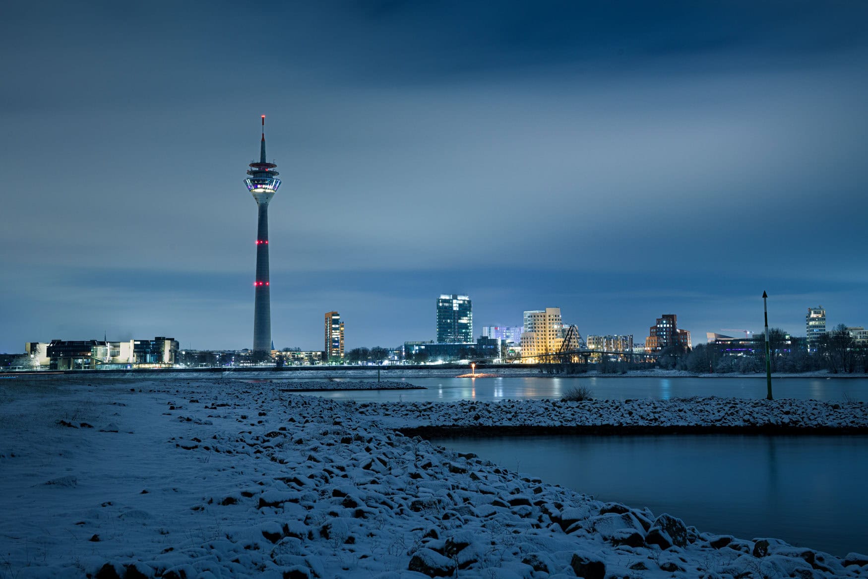 Verschneite Rheinuferwiesen mit Düsseldorfer Rheinturm und Skyline bei Nacht