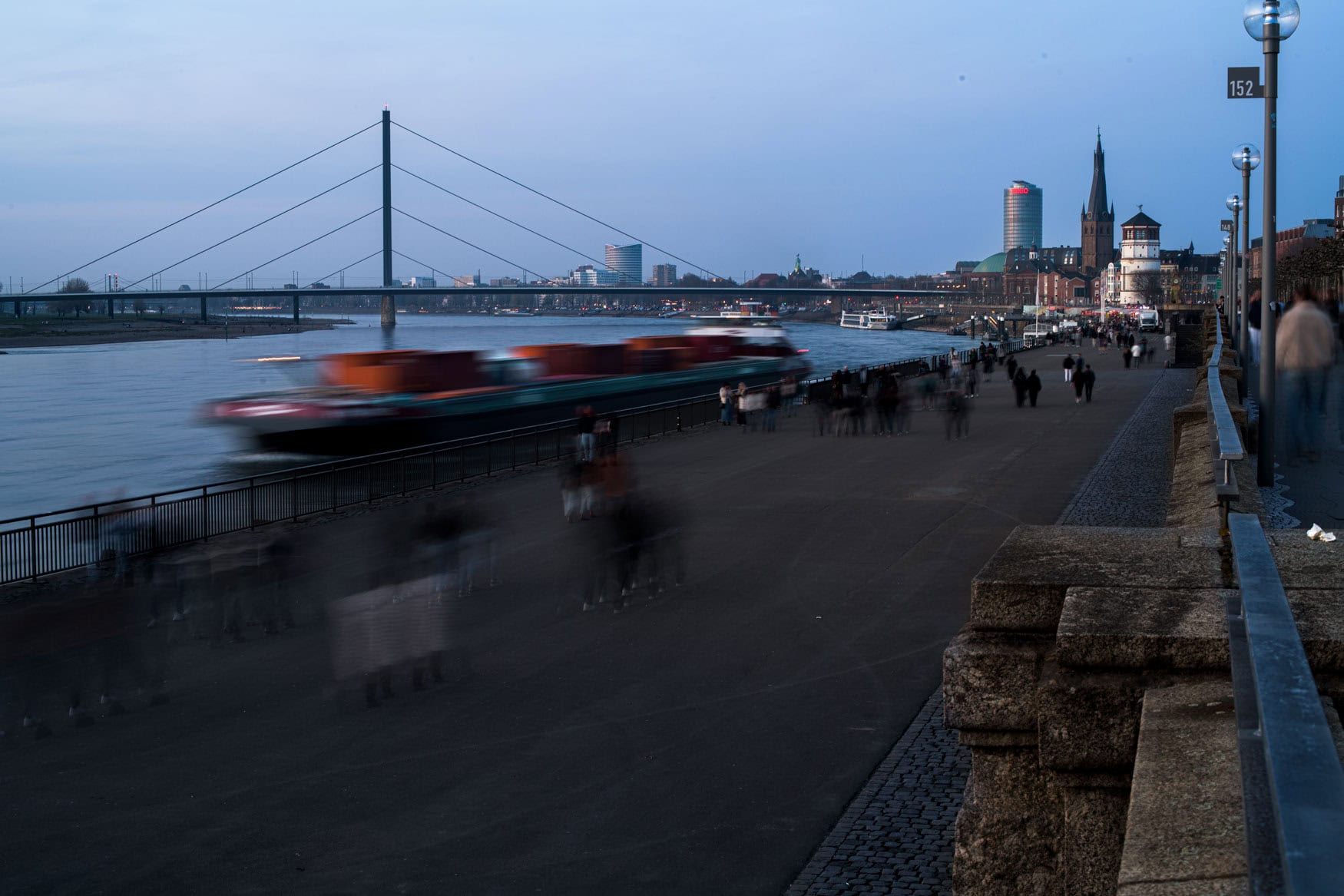 Langzeitbelichtung der Düsseldorfer Rheinuferpromenade mit Frachtschiff bei Abendlicht