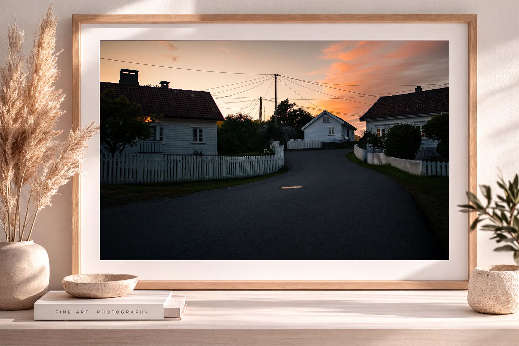 Fotografie einer leeren Dorfstraße mit weißen Holzhäusern und Lattenzäunen bei warmem Sonnenuntergang im gerahmten Wandbild