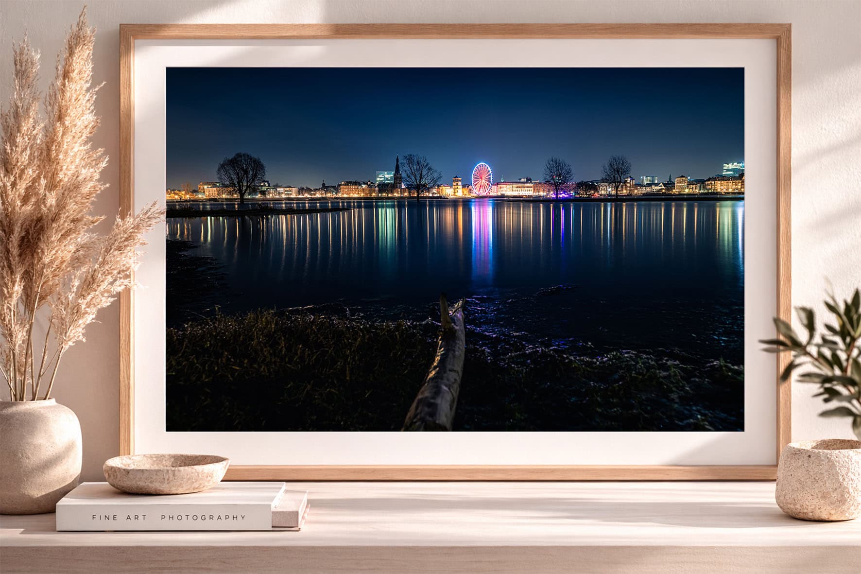 Fotografie einer nächtlichen Stadt am Wasser mit beleuchtetem Riesenrad, gespiegelt im ruhigen Fluss, in hellem Holzrahmen auf Sideboard.