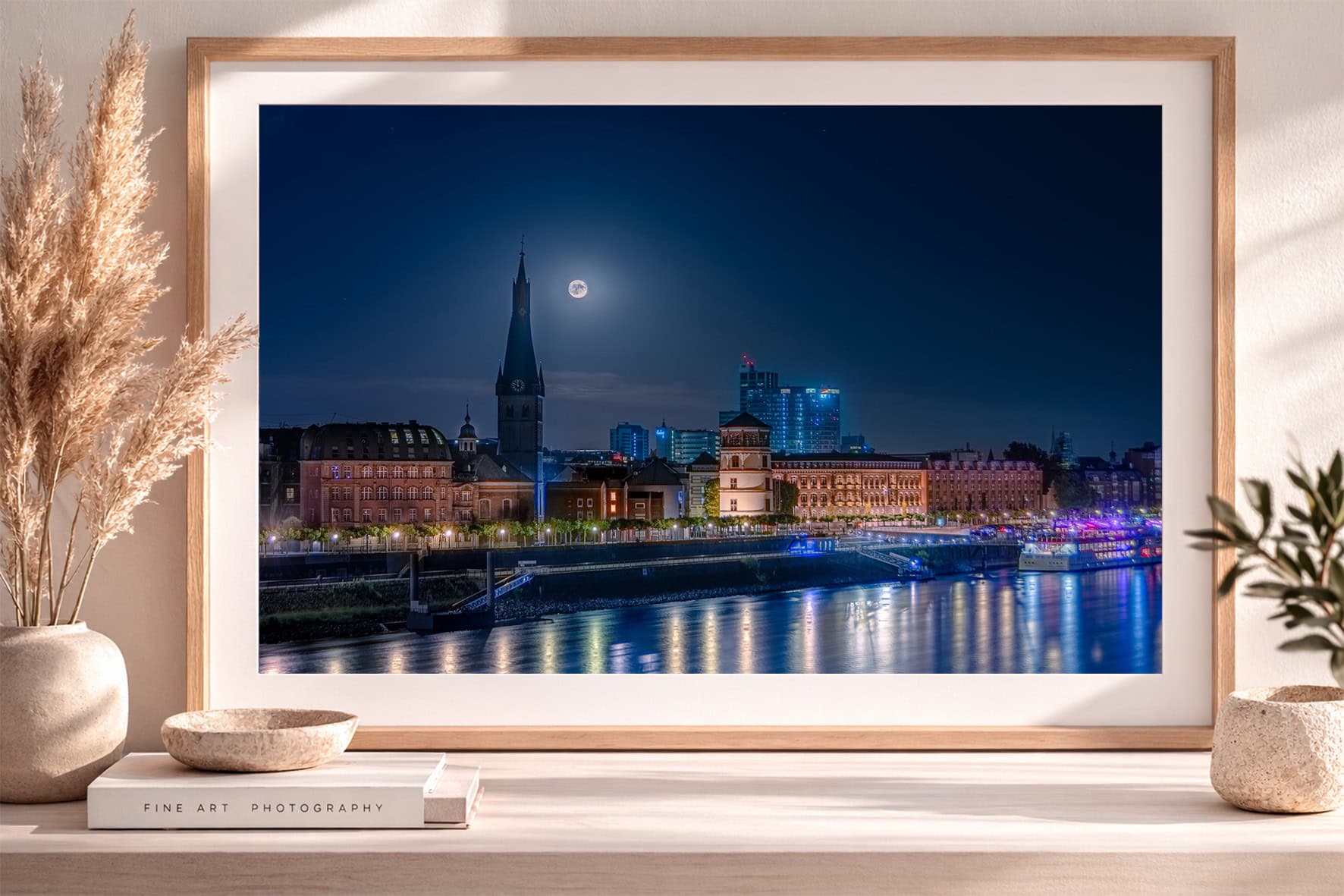 Großer gerahmter Kunstdruck einer nächtlichen Stadtansicht am Fluss mit Vollmond, beleuchteten Gebäuden und Spiegelungen, auf Sideboard dekoriert.