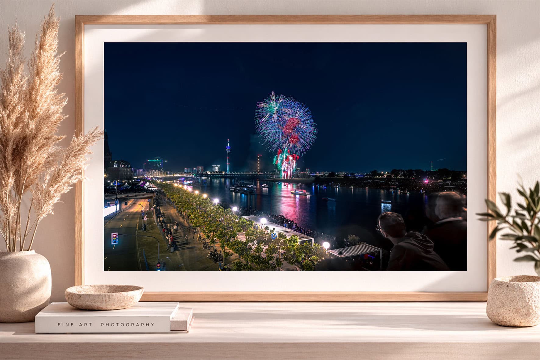 Fotokunst mit Feuerwerk über einer beleuchteten Uferpromenade am Fluss bei Nacht in hellem Holzrahmen auf Sideboard.