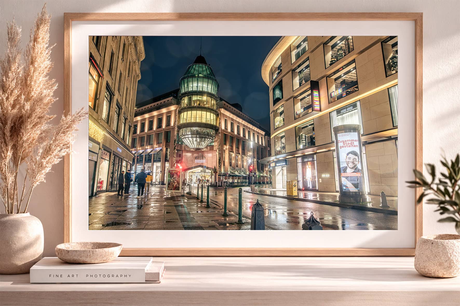 Fotografie einer beleuchteten Einkaufsstraße bei Nacht mit Glasbau, nassem Pflaster und Passanten in einem gerahmten Wandbild im Interior.
