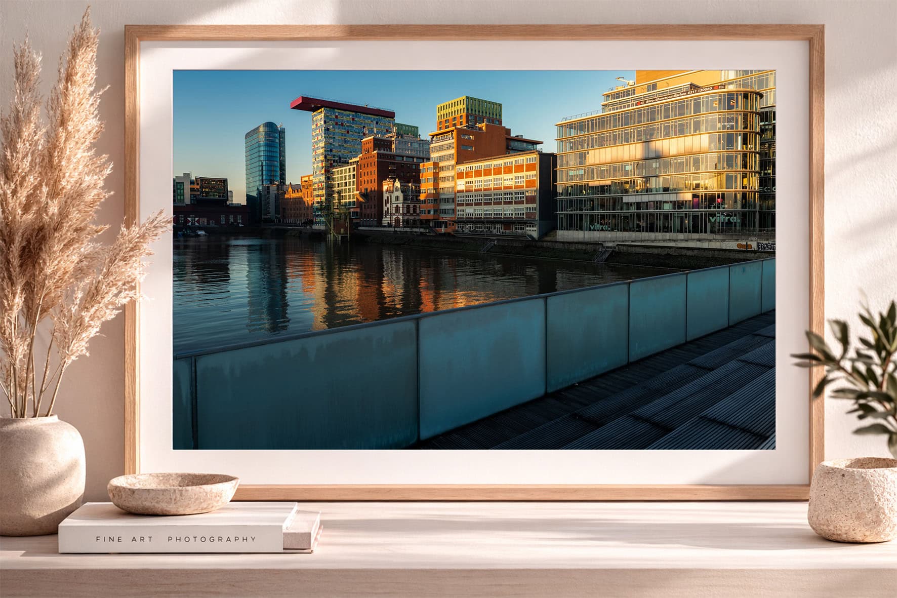 Fotografie moderner Bürogebäude am Fluss, reflektiert im Wasser, gesehen von einer Uferpromenade bei warmem Abendlicht im Rahmen.