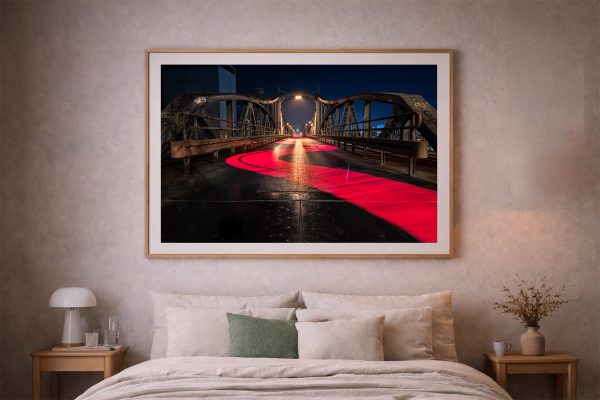 Fotografie einer beleuchteten Stahlbrücke bei Nacht mit geschwungener roter Lichtspur, gerahmt über einem Bett in einem dezenten Schlafzimmer