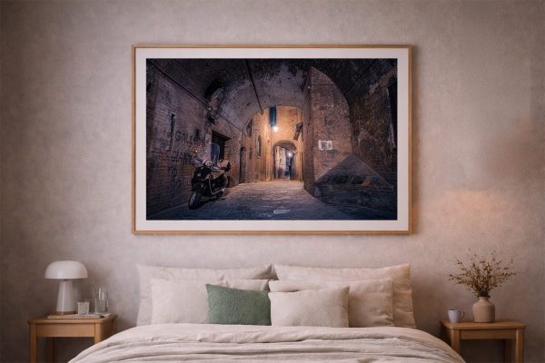 Fotografie einer beleuchteten Altstadtgasse mit Vespa unter Backsteinbögen, als gerahmtes Wandbild über einem Bett im Schlafzimmer
