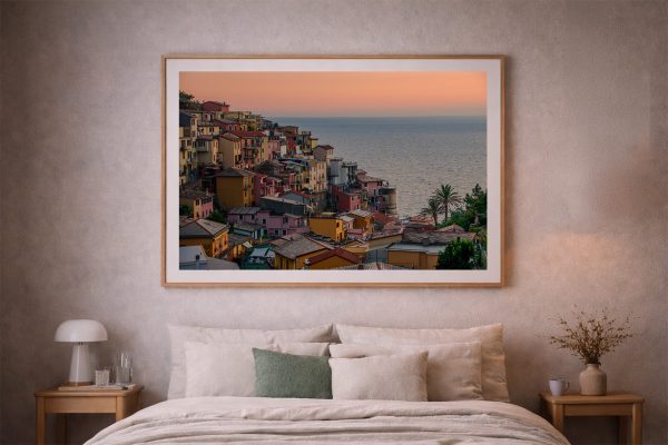 Fotografie einer bunten Küstenstadt vor ruhigem Meer im Sonnenuntergang, gerahmt über einem beige eingerichteten Bett im Schlafzimmer.