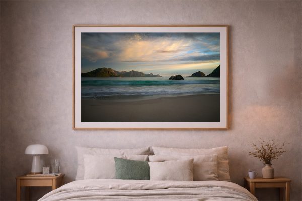 Großer gerahmter Küstenprint mit Bergen und sanften Wellen über einem Bett in einem ruhigen Schlafzimmer