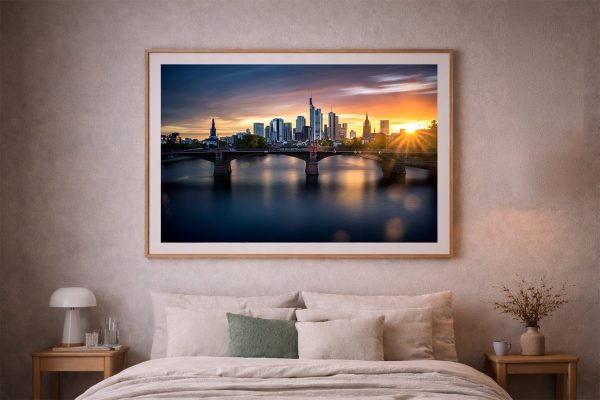 Großer gerahmter Kunstdruck einer Skyline mit Brücke über ruhigem Fluss bei warmem Sonnenuntergang über einem Bett im Schlafzimmer.