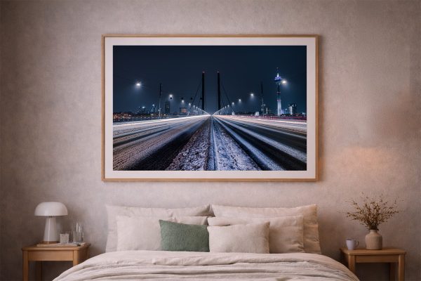 Fotografie einer beleuchteten Stadtbrücke bei Nacht mit verschneiter Fahrbahn und Lichtspuren, gerahmt über einem Bett im gemütlichen Schlafzimmer