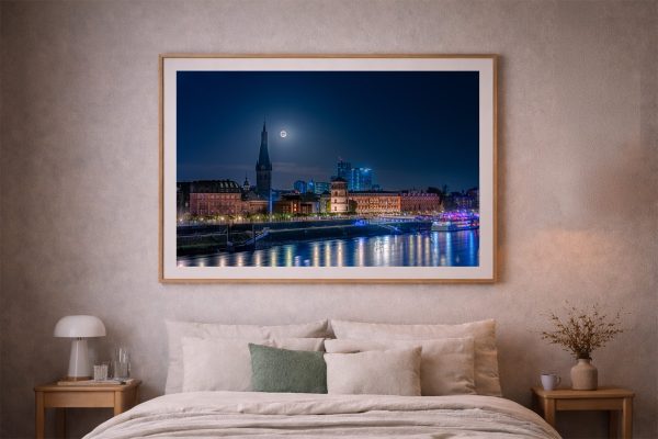 Fotografie einer nächtlichen Stadtansicht mit Fluss, beleuchteten Gebäuden und Vollmond, gerahmt über einem Bett im gemütlichen Schlafzimmer