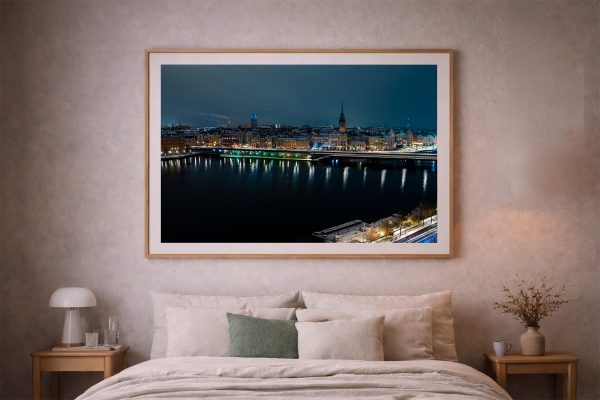 Großes gerahmtes Foto einer erleuchteten Stadt bei Nacht über dunklem Wasser, über einem Bett in einem dezent eingerichteten Schlafzimmer.