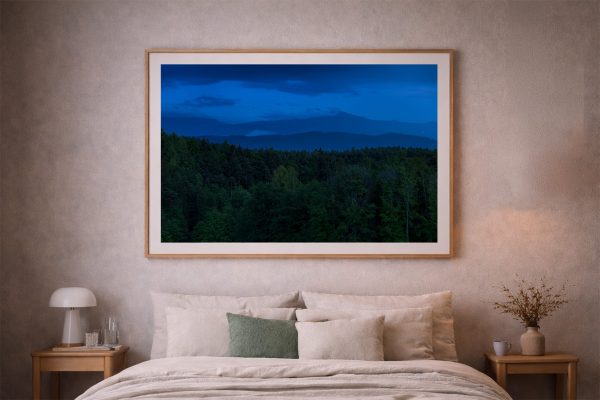 Großer gerahmter Kunstdruck mit dunkler Waldlandschaft und blauen Bergen hängt über einem beigefarbenen Bett im Schlafzimmer.