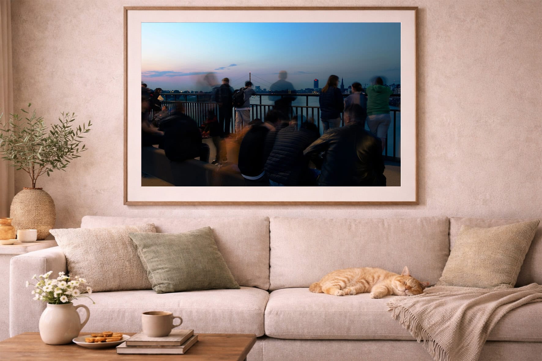 Fotografie mit unscharfen Menschen an einer Uferpromenade bei Abenddämmerung, gerahmt über einem beigen Sofa im gemütlichen Wohnzimmer