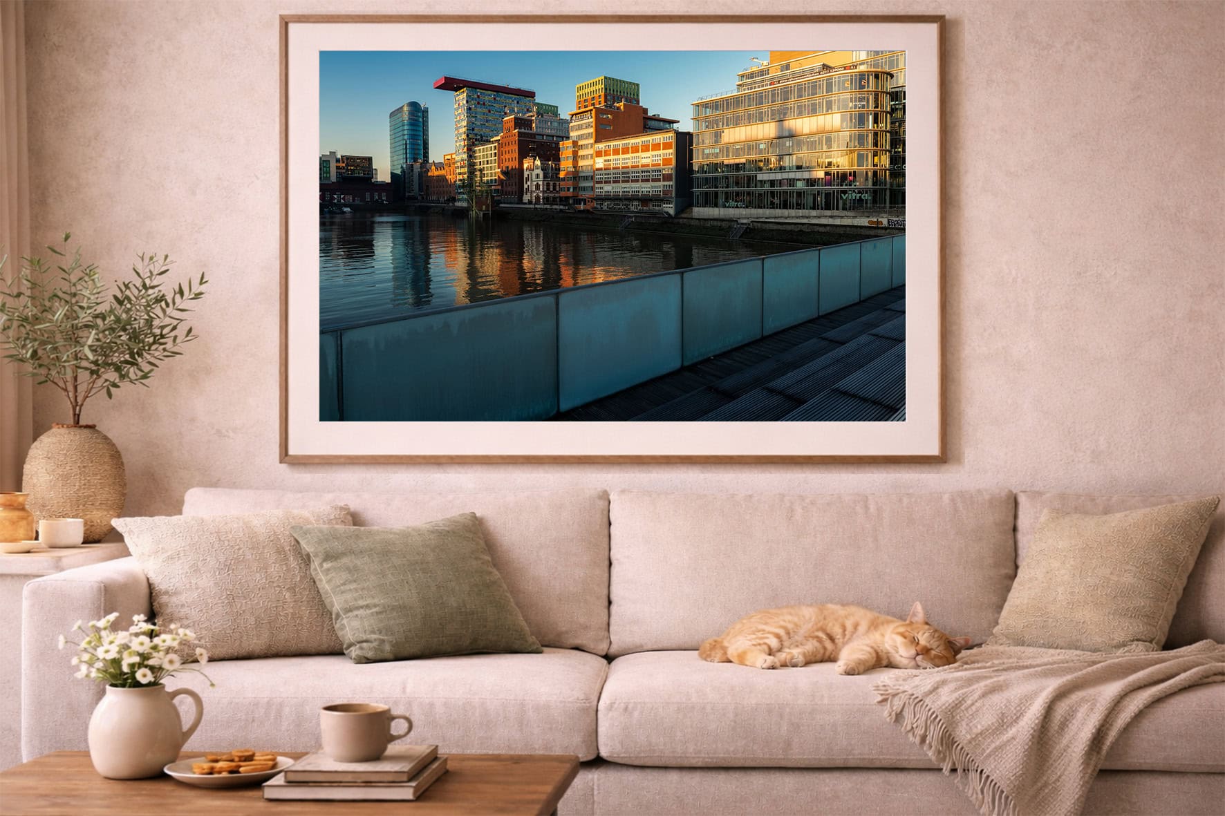 Fotografie moderner Gebäude am Fluss im Abendlicht, gespiegelt im Wasser, als gerahmtes Wandbild über beigem Sofa mit Katze