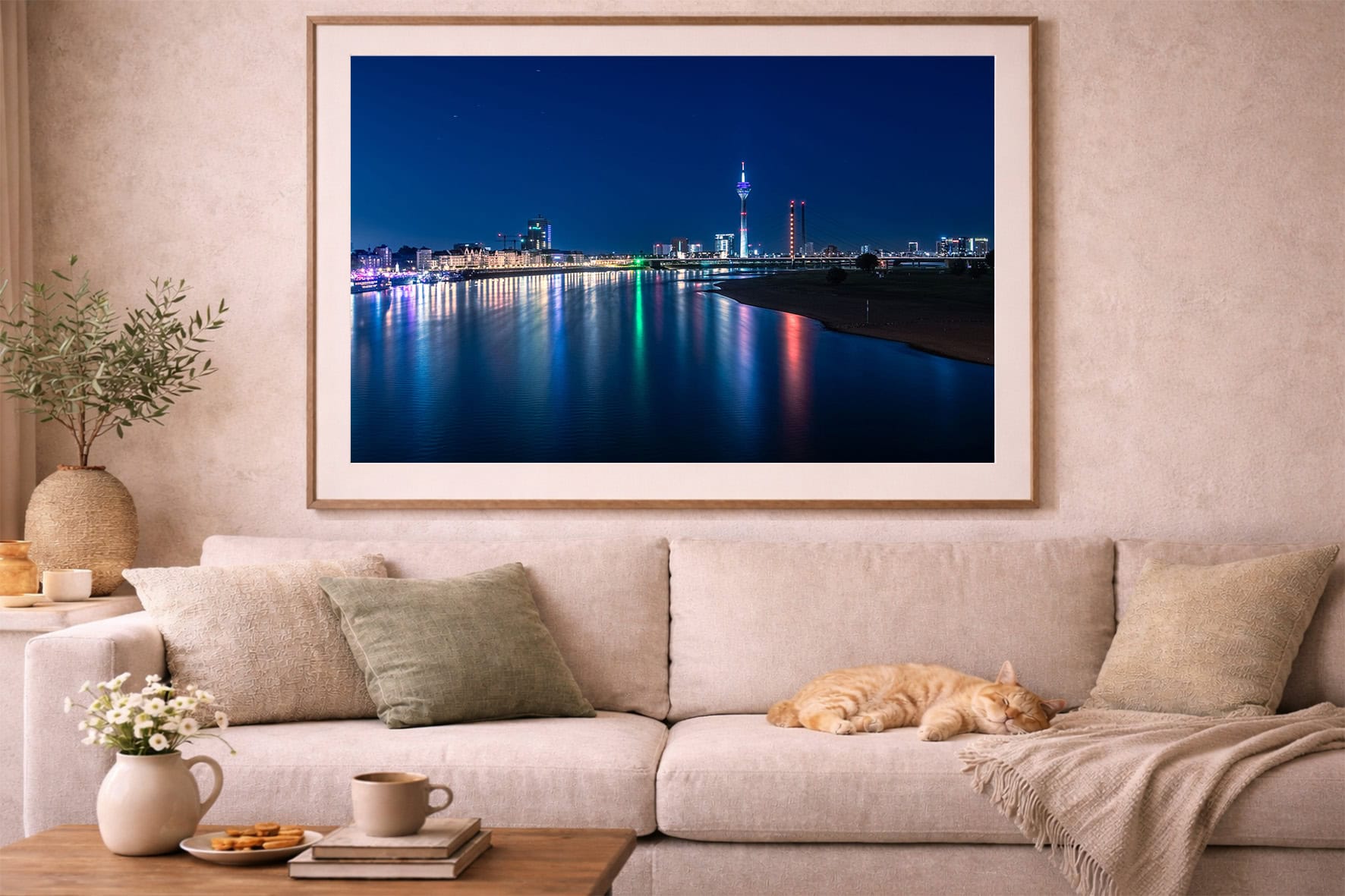 Fotografie einer nächtlichen Stadt am Fluss mit bunten Lichtern im Rahmen über einem beigen Sofa mit schlafender Katze.