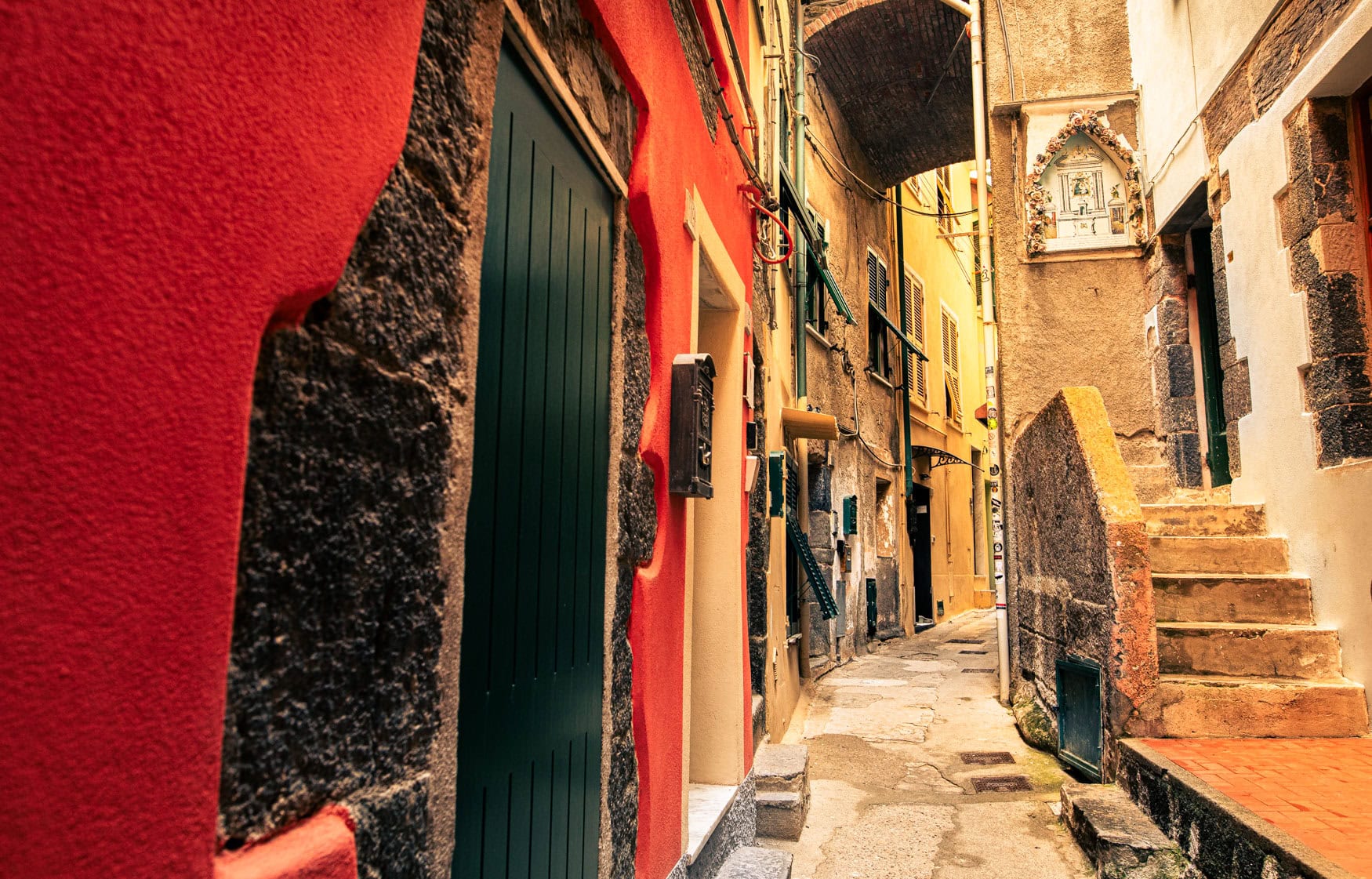 Schmale Gasse in Cinque Terre mit roter Fassade, grünen Türen, Treppe und dicht stehenden Häusern