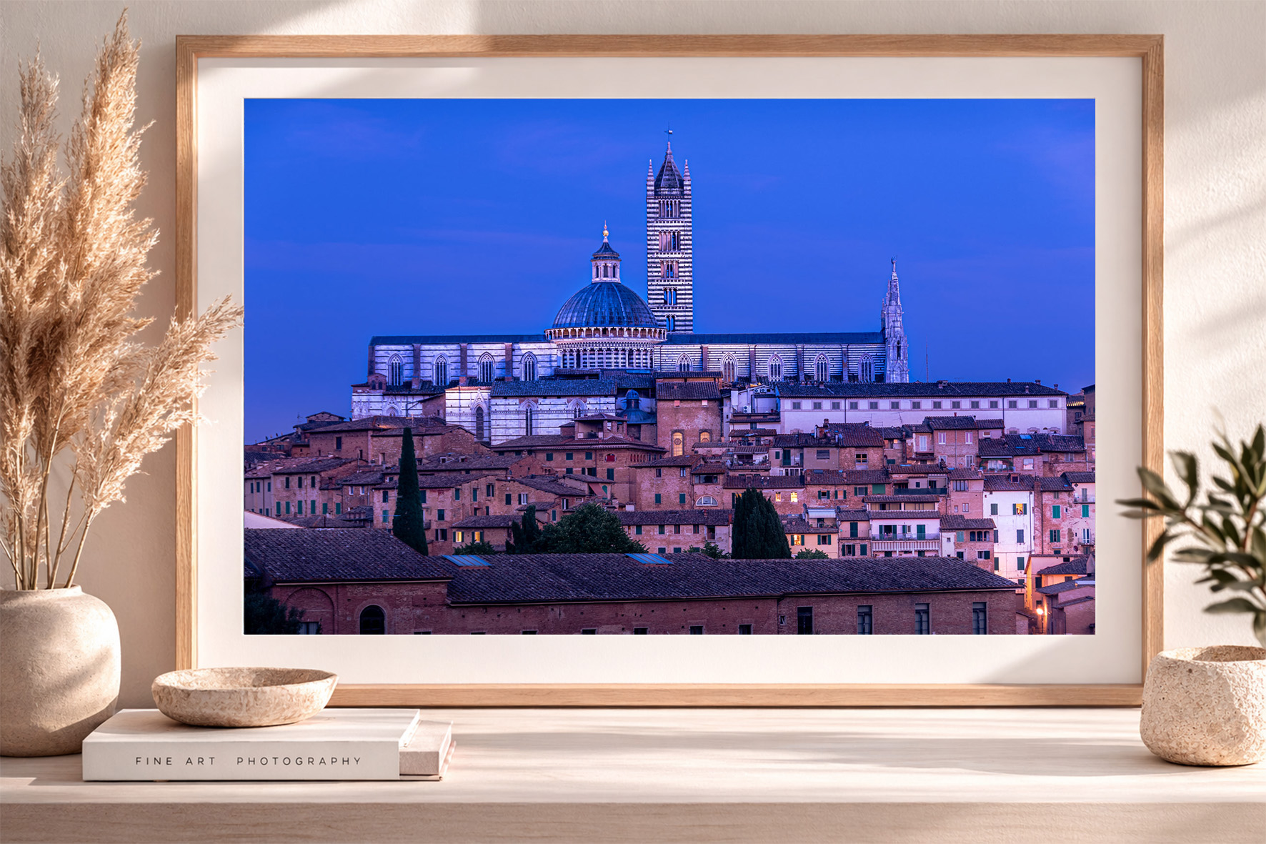 Fine-Art-Fotografie einer beleuchteten Altstadt mit großer Kathedrale bei blauem Abendhimmel in hellem Holzrahmen auf Sideboard.