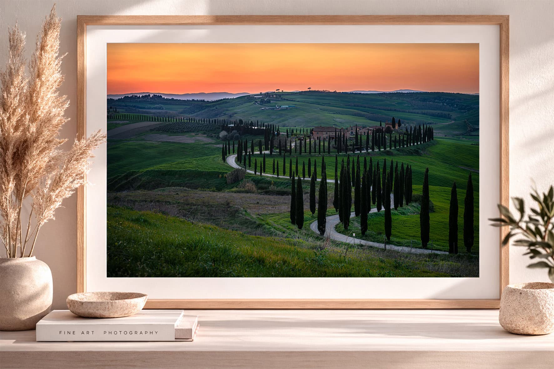 Gerahmtes Landschaftsfoto mit kurviger Zypressenallee durch grüne Hügel unter orangefarbenem Abendhimmel auf Sideboard im modernen Interior.