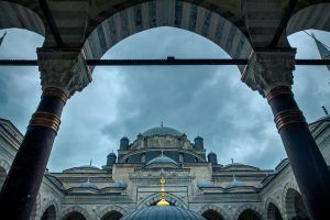 Blick durch einen Arkadenbogen auf eine Moschee in Istanbul mit Kuppeln, Säulen und wolkigem Himmel