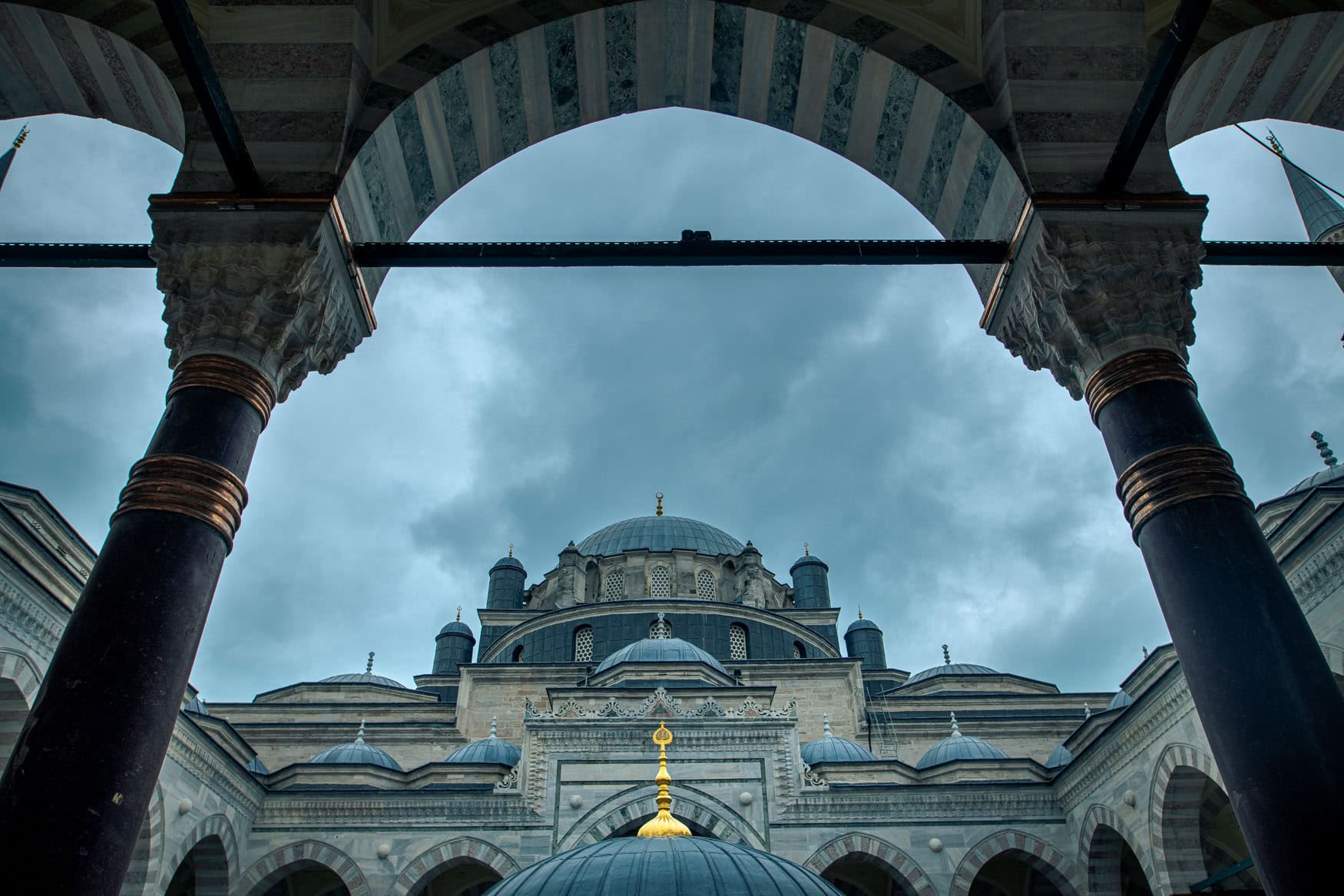 Blick durch einen Arkadenbogen auf eine Moschee in Istanbul mit Kuppeln, Säulen und wolkigem Himmel