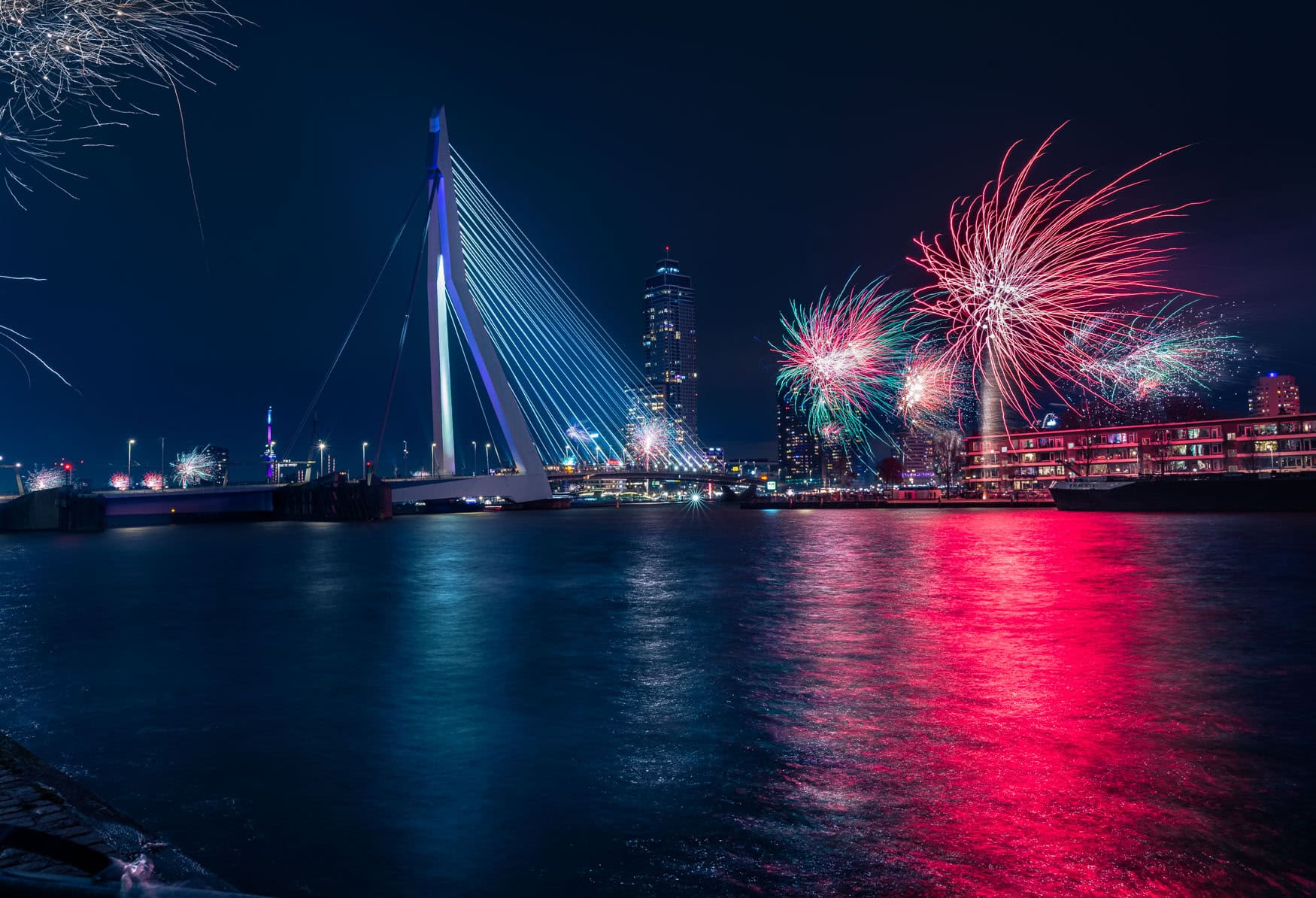 Feuerwerk über der Erasmusbrücke in Rotterdam bei Nacht mit farbigen Spiegelungen auf dem Wasser