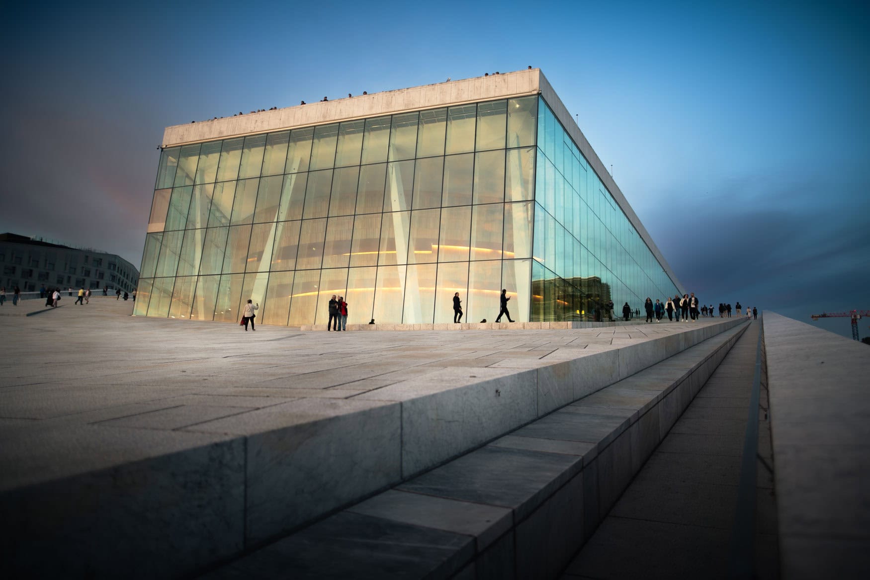 Gläserne Architektur des Osloer Opernhauses in der Abenddämmerung mit Menschen auf der hellen Steinfläche