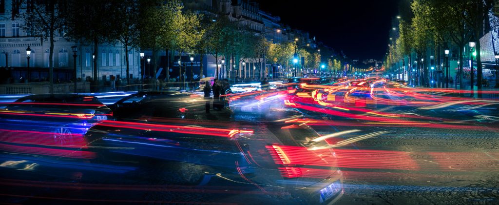 Nächtlicher Kreisverkehr am Arc de Triomphe in Paris mit roten und weißen Lichtspuren und beleuchteten Bäumen