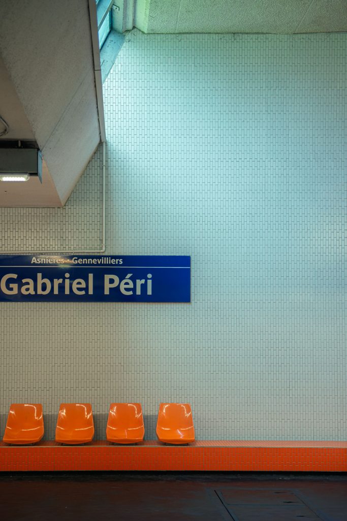 Metrostation Gabriel Péri in Paris mit blauem Stationsschild, heller Kachelwand und vier orangenen Sitzen