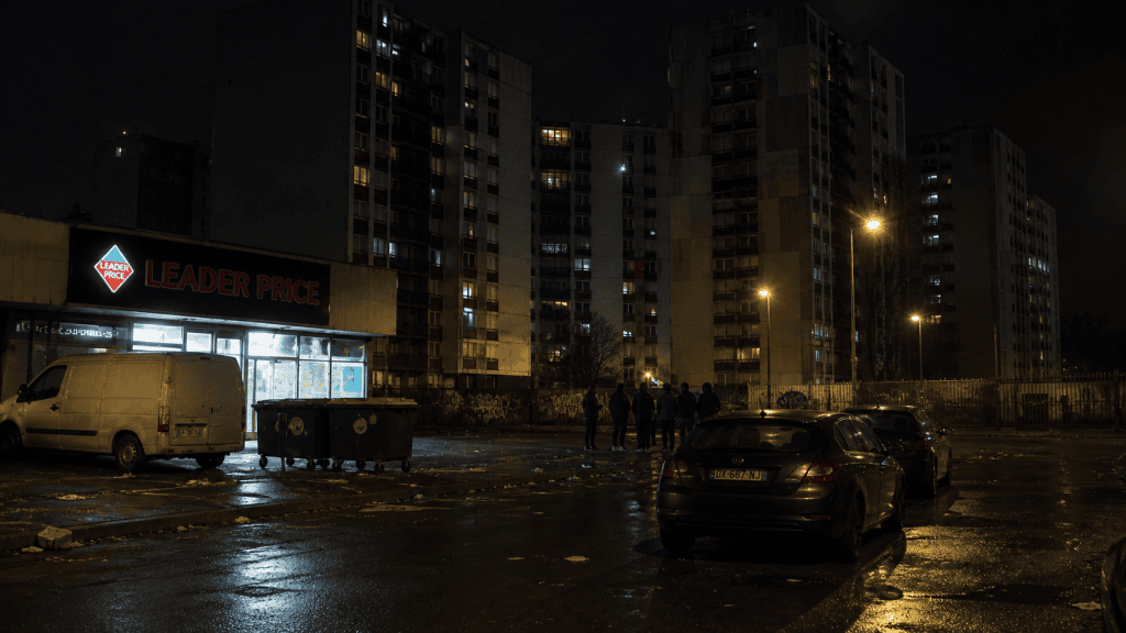 Nachtaufnahme in Saint-Denis mit Wohnhochhäusern, beleuchtetem Laden, nasser Straße und parkenden Autos
