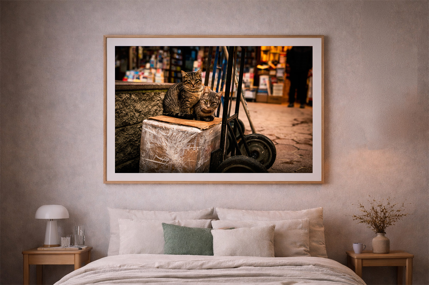 Fotografie von zwei getigerten Katzen auf einer Folienkiste neben Sackkarre, in warm beleuchteter Stadtgasse, über einem Bett gerahmt.