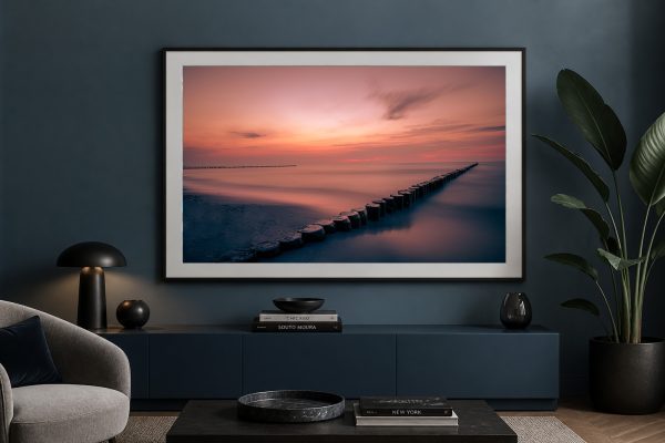 Großes gerahmtes Foto eines ruhigen Meeres mit Holzpfählen im Sonnenuntergang über einem dunkelblauen Sideboard im Wohnzimmer